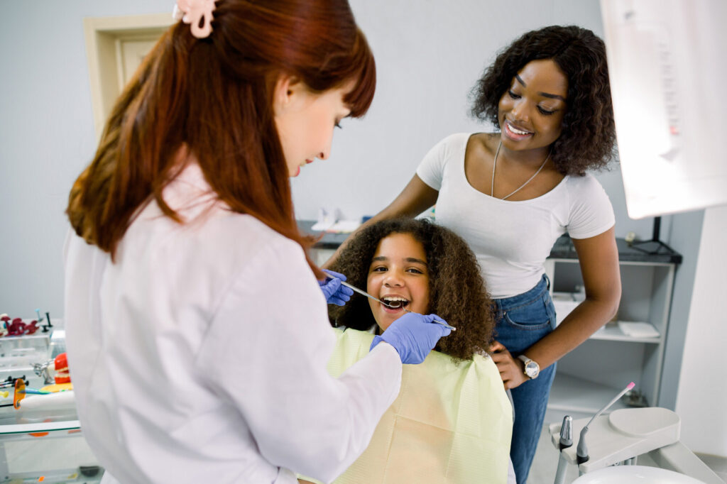 Young,African,Woman,Mom,Supporting,Her,Little,Daughter,At,Dentist's
